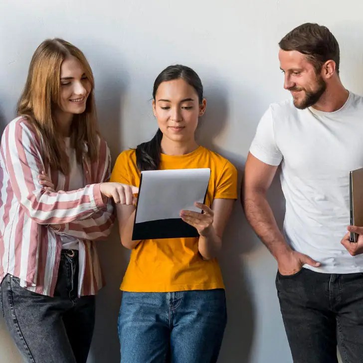 Drei Menschen, zwei Frauen und ein Mann, stehen an einer weißen Wand gelehnt, die mittlere Person hält ein Klemmbrett mit einem Blatt Papier, auf das alle drei Personen interessiert schauen.